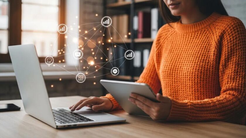 Kvinna i orange tröja arbetar vid laptop och surfplatta med digitala ikoner som symboliserar data, e-post och nätverk – ett visuellt koncept för Marketing as a Service (MaaS).